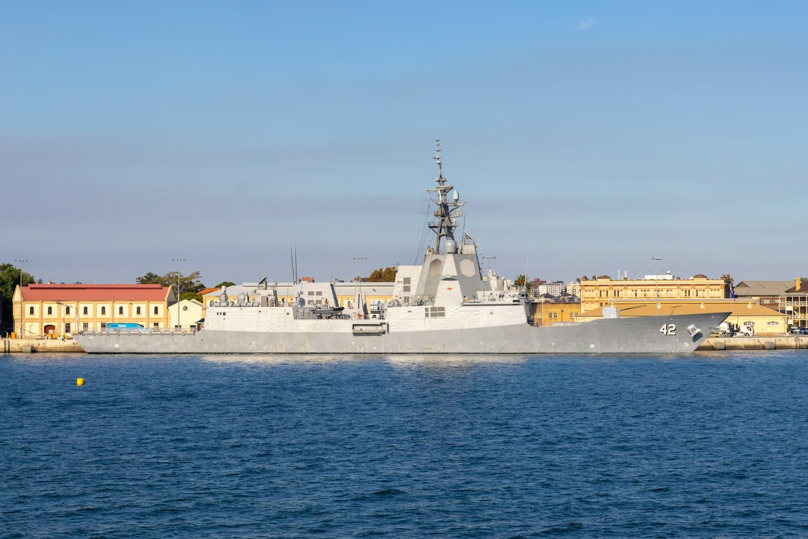 A modern grey warship alongside, with waterfront buildings behind